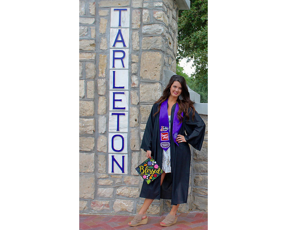 Shelbi Tidwell wearing her purple graduation gown, holding her graduation cap next to a Tarleton State University sign. 